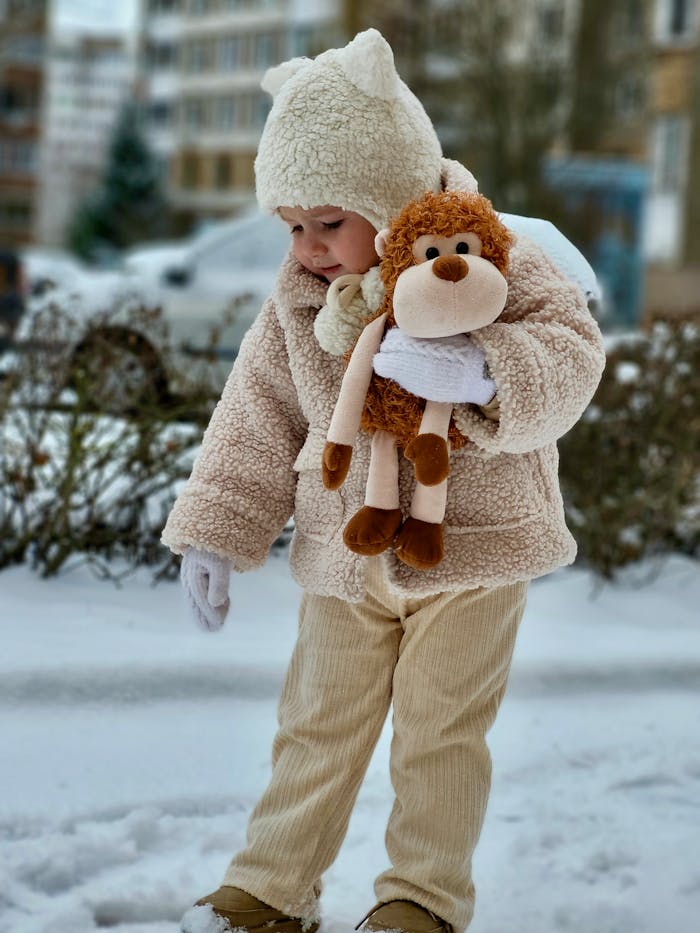 Adorable child in warm winter clothing holding a plush toy in a snowy urban setting.