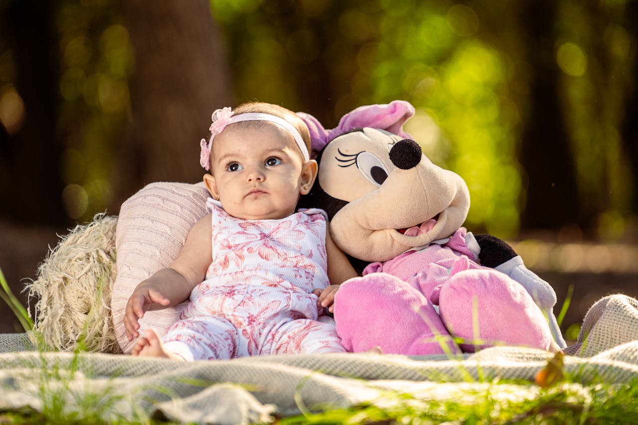 Charming portrait of a baby girl with a stuffed Minnie Mouse toy in a serene outdoor setting.
