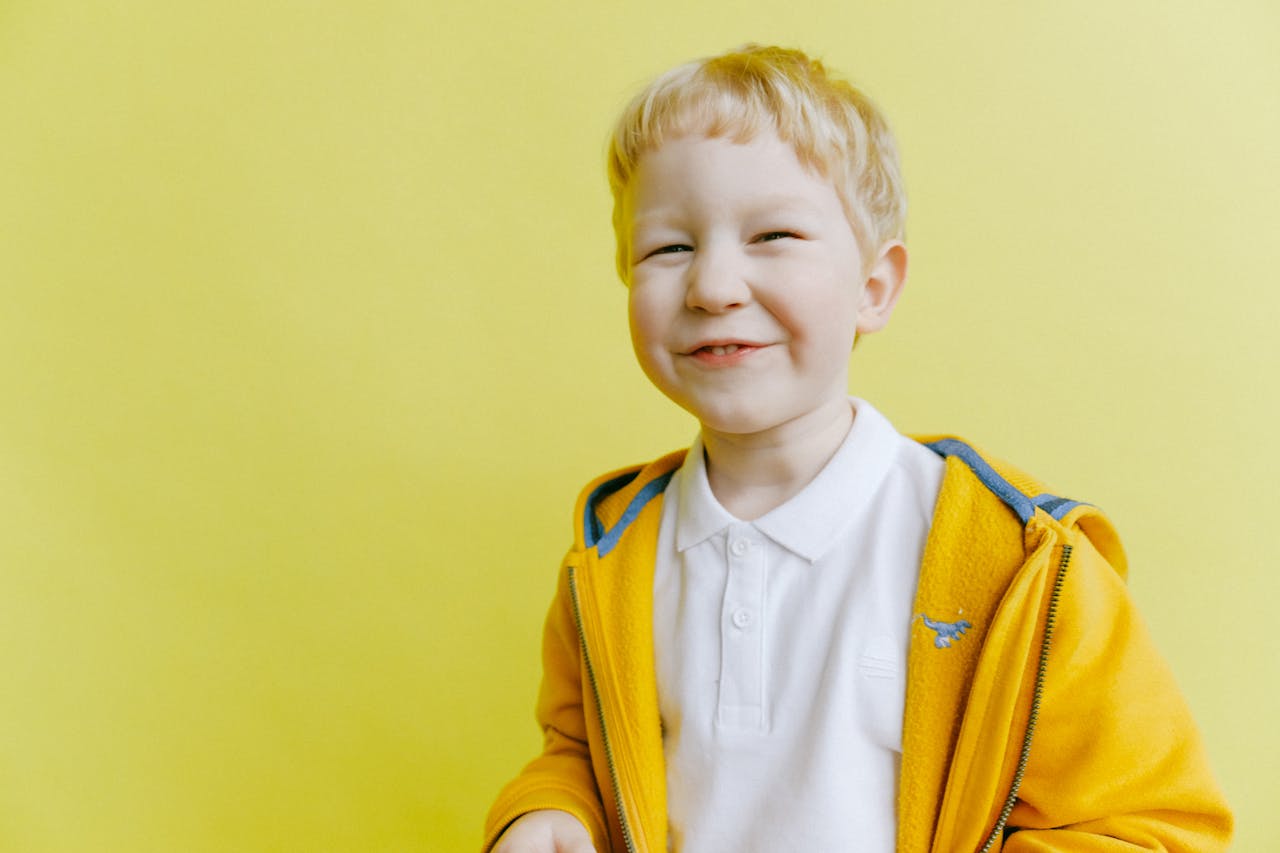 Cheerful young boy with blond hair smiling in a studio setting against a vibrant yellow background.