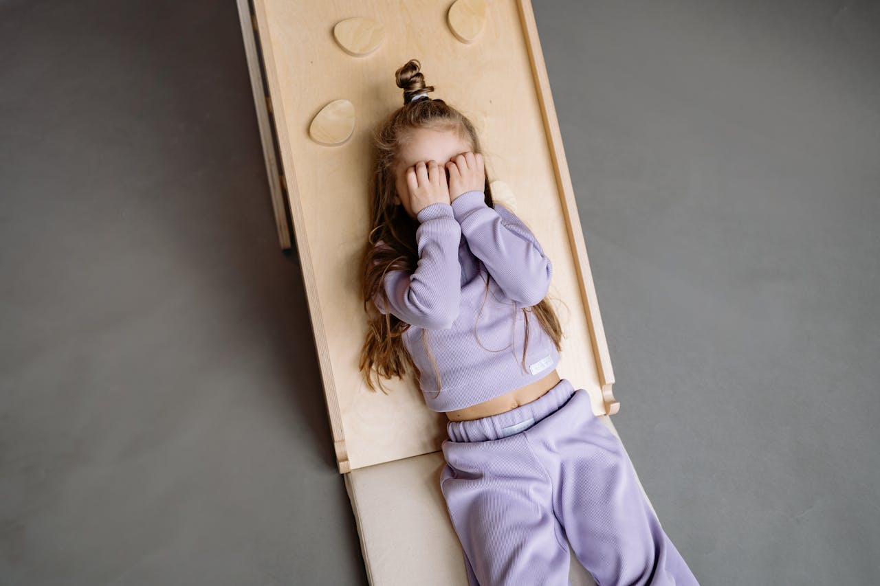 Cute girl in purple outfit covering face while lying on a wooden slide indoors.